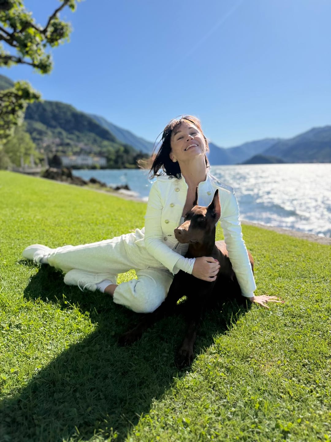 Masha sitting on the grass beside a Doberman by Lake Como, with the lake and mountains behind them.