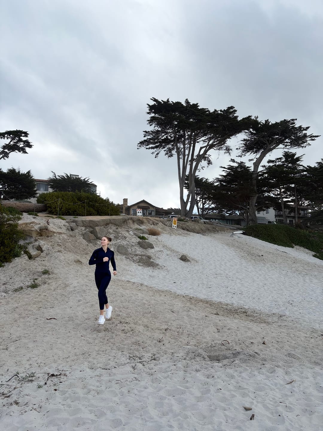 A single person running across an empty beach below dunes and cypress trees under a grey sky.