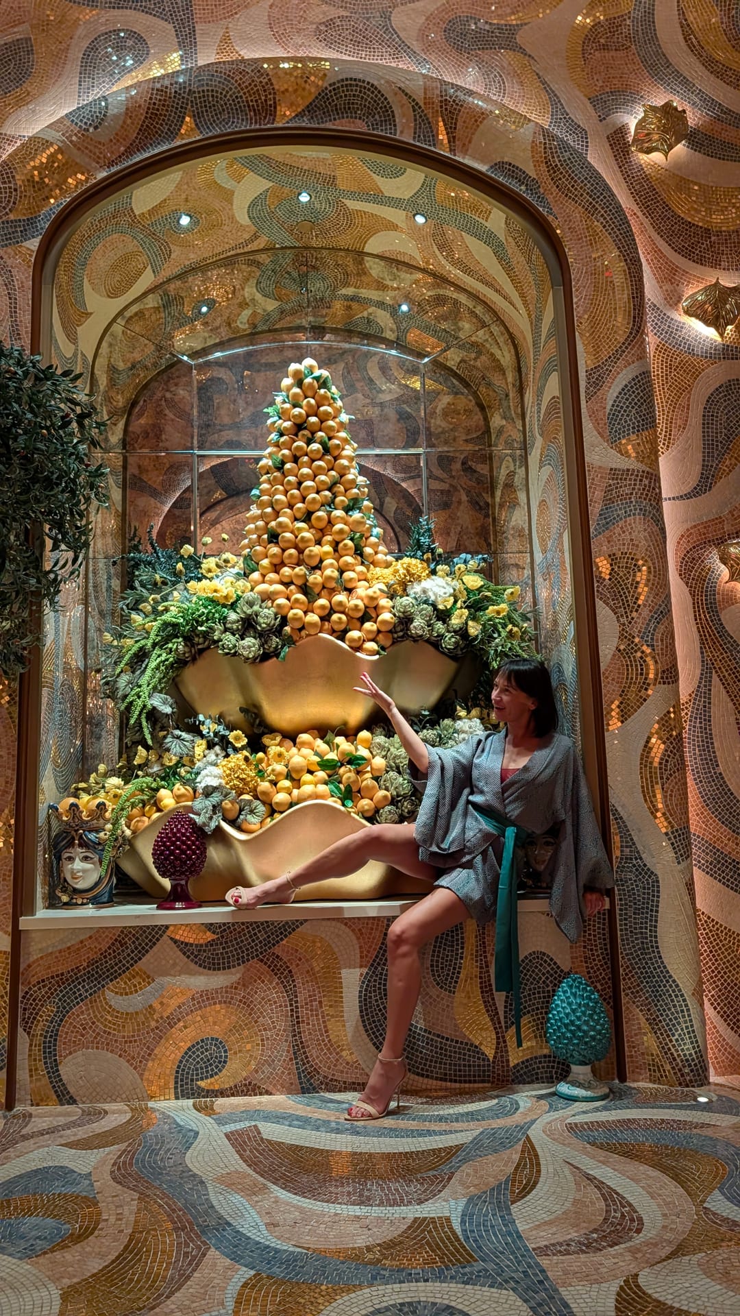 Masha seated with one leg extended in front of a gold-and-citrus display against a mosaic wall.